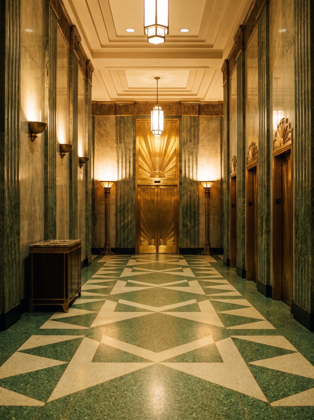 The elevator hall: fluted verd-antique marble walls, brass elevator doors chased with a sunburst, torchère lamps, and a green-and-cream terrazzo floor.