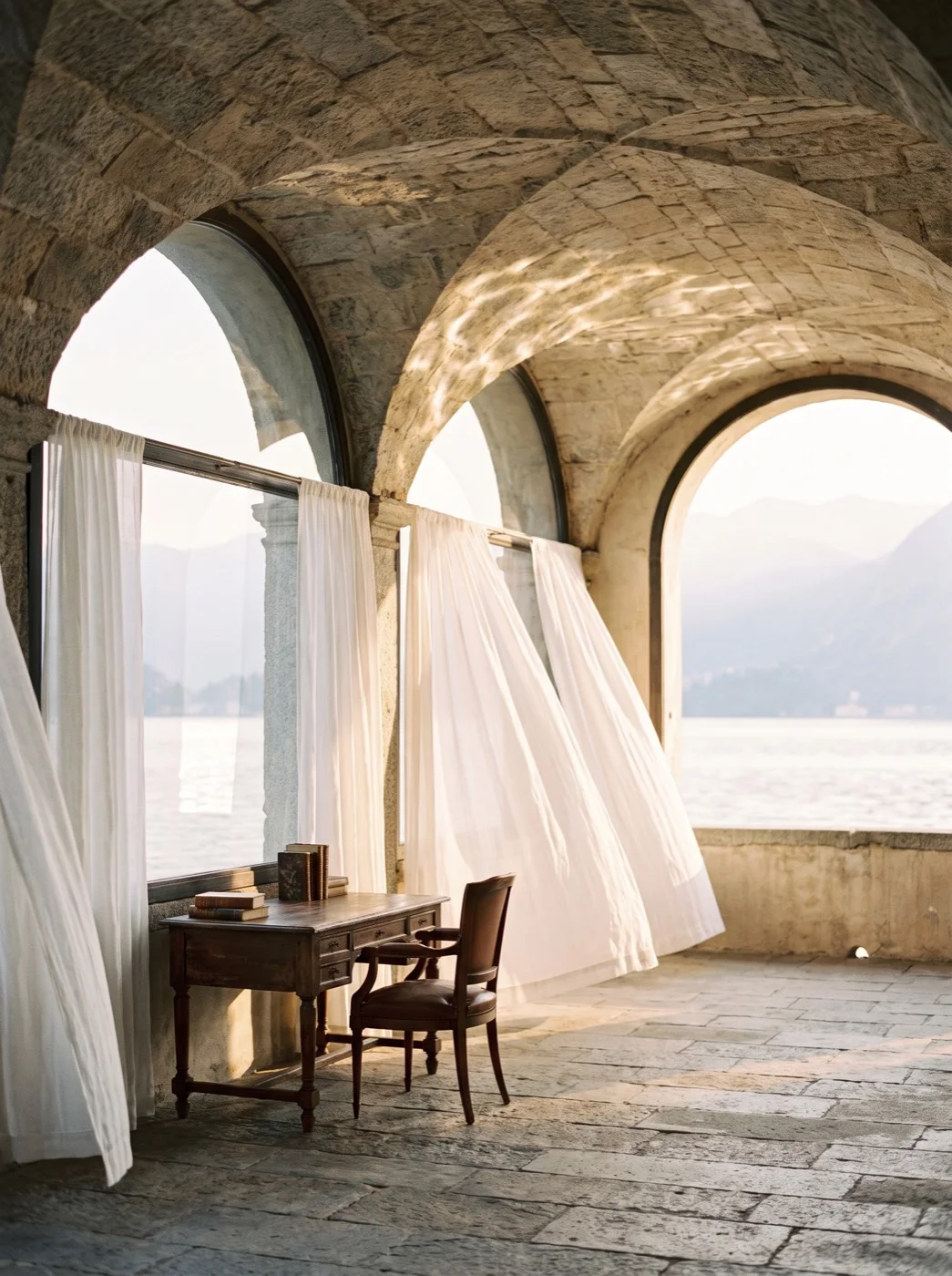 The villa loggia: vaulted stone bays open to the lake, white linen curtains lifting in the wind, a writing desk and single chair on worn flagstones.