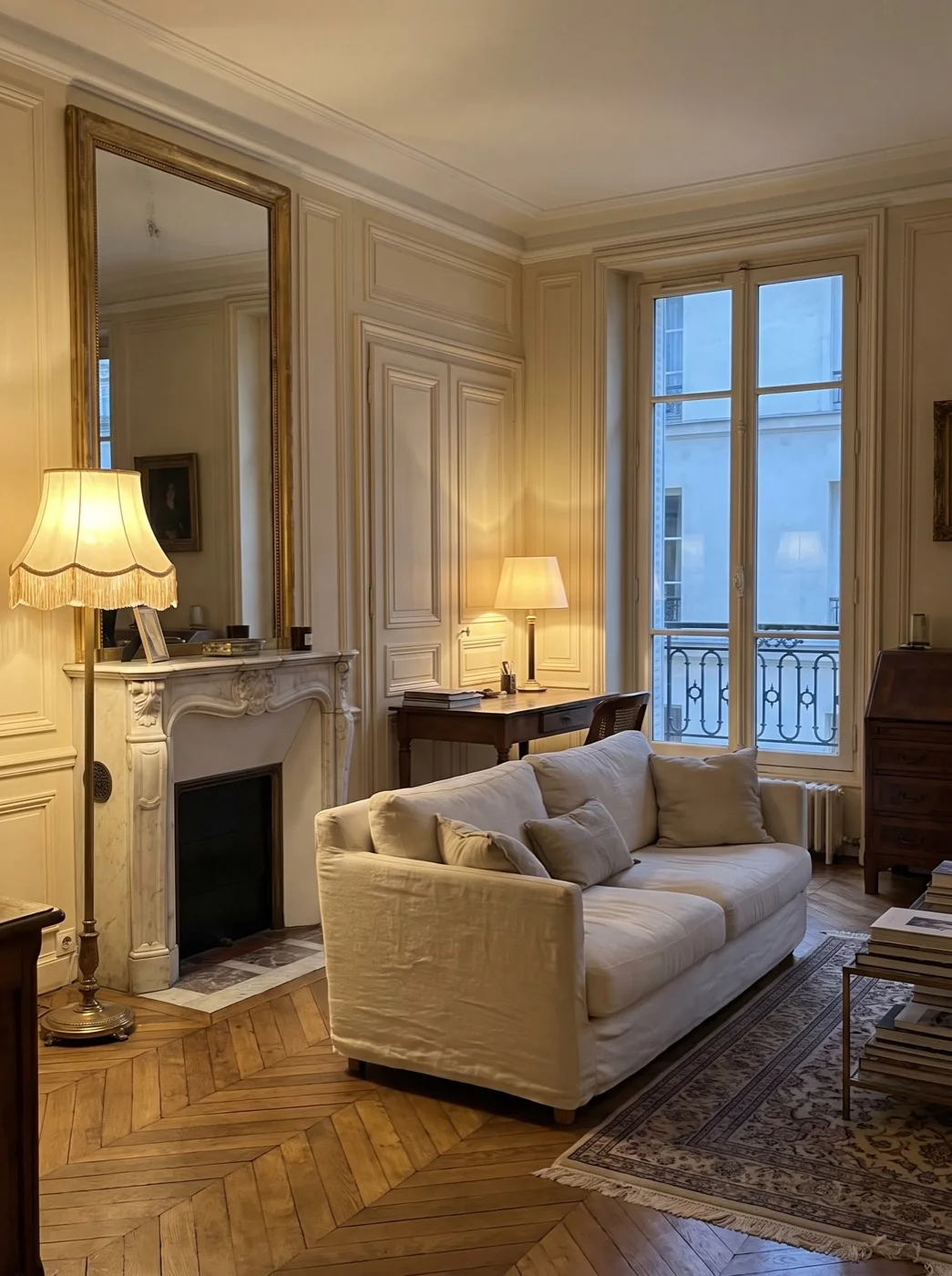 The Paris salon at evening: chevron oak parquet, a carved marble chimney under a gilt mirror, a linen sofa, two lamps already lit.