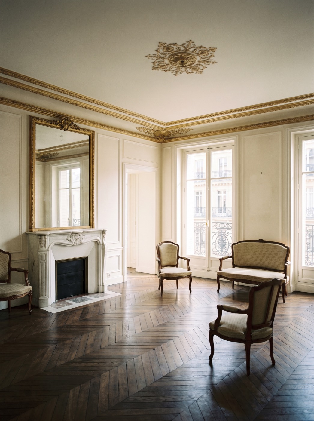 The salon at 21 rue de Seine: chevron oak parquet, white marble fireplace with gilt trumeau mirror, gilt cornice and ceiling rose, tall windows onto the street.