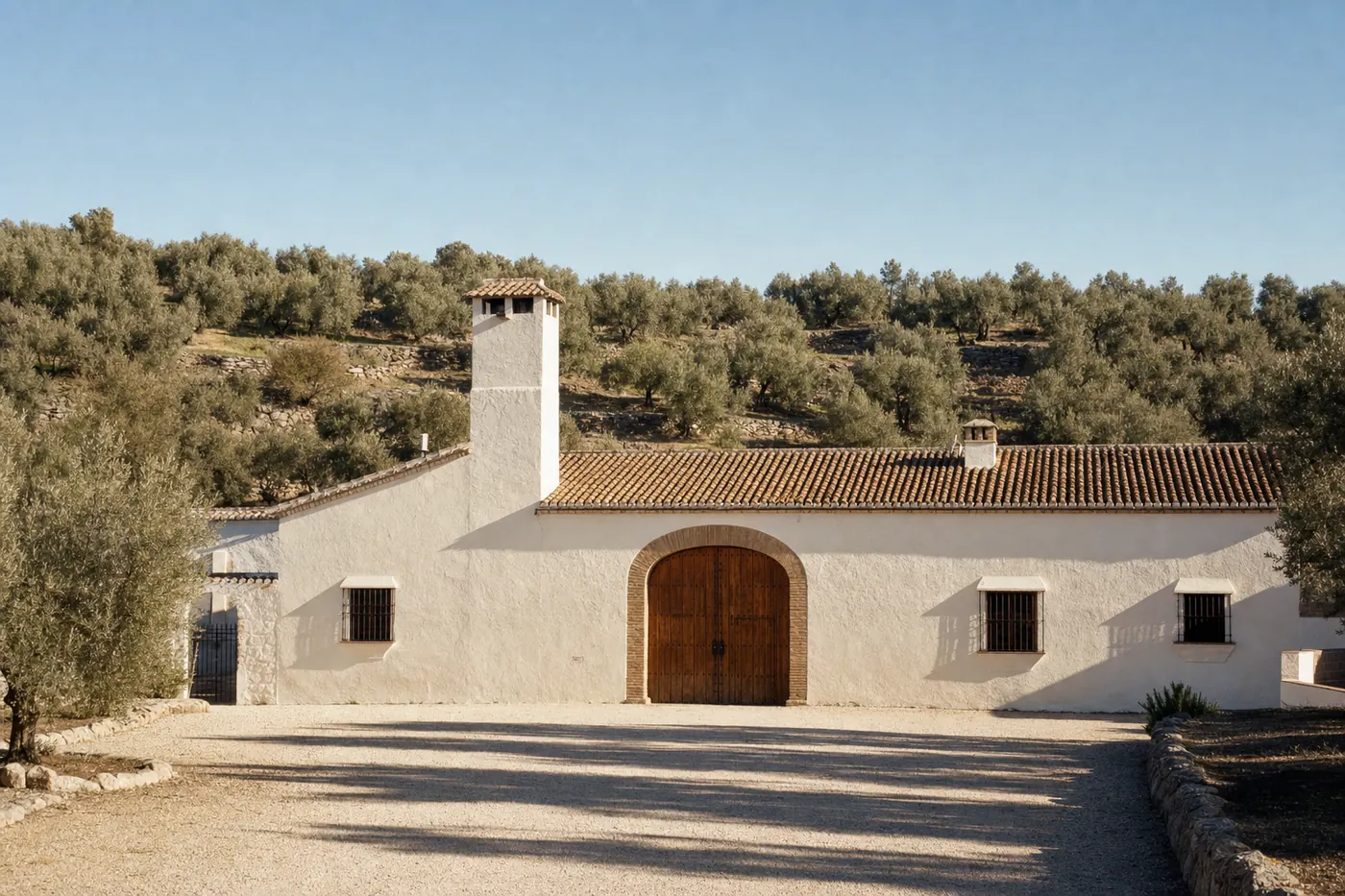 El Molino de las Aguas: a long whitewashed olive-mill cortijo with a square chimney tower and an arched chestnut mill door, olive terraces rising behind it in morning light.